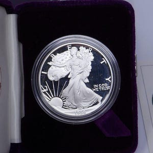 May include: A silver American Eagle coin from 1986, displayed in a protective case with a purple velvet interior. The coin features Lady Liberty walking with the sun rising behind her, and the words "IN GOD WE TRUST". The case has the words "AMERICAN EAGLE" on the lid.
