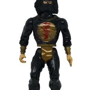 Puede incluir: Figura de acción negra y dorada de un guerrero ninja. La figura tiene una máscara negra, pelo negro y un atuendo negro y dorado. El pecho presenta un diseño rojo y dorado. La figura está de pie con los brazos a los lados.