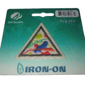 Pode incluir: Um patch Try-Its das Girl Scouts em um suporte verde-azulado e branco. O patch triangular apresenta formas e linhas bordadas coloridas. O verso diz "IRON-ON" e inclui o logotipo das Girl Scouts.