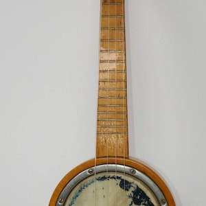 May include: Vintage banjo with a wooden neck and body. The round body features a painted design with blue and white elements. The instrument has strings and tuning pegs, showcasing a classic design.