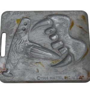 May include: A vintage, rectangular metal mold with a handle, featuring a dinosaur claw and a bird-like figure. The mold is gray with some yellow and brown discoloration. The text "©1986 MATTEL, INC. U.S.A" is visible.