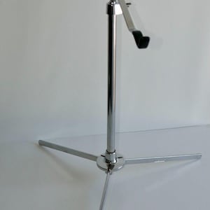 May include: A chrome-plated music stand with a tripod base and adjustable height. The stand features a rectangular plate with the brand name and three angled arms with black rubber supports for holding sheet music. The stand is designed for musicians.