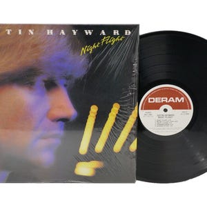 May include: A vinyl record album titled "Night Flight" by Justin Hayward. The album cover features a portrait of a man with blonde hair and a dark background. The record itself is black with a white and red label.