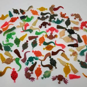 May include: A collection of small, colorful plastic toys in various shapes, including frogs, horns, and abstract forms. The toys are in shades of red, green, yellow, orange, and brown, scattered on a white surface.