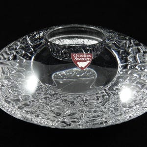 May include: Clear glass bowl with a textured, patterned exterior and a central, raised circular holder. The bowl features the "Orrefors Sweden" logo. The design has a textured, grid-like pattern. The bowl is on a black background.