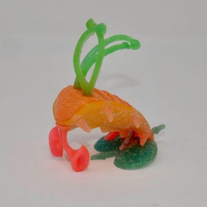 May include: A small, multi-colored 3D printed figurine featuring an orange, textured, curved body with two bright green, thin, arching antennae on top. It has two pink, claw-like appendages at the front and rests on a green, speckled base.