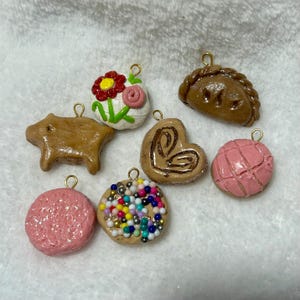 May include: A set of eight handmade clay charms, each depicting a different Mexican pastry or sweet treat. The charms are brown, pink, and white with colorful details and have gold-colored loops for hanging.