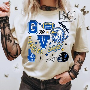 May include: Cream-coloured t-shirt featuring blue and yellow graphics. The design incorporates a football, an eagle, the letters "G" and "V", the word "EAGLES", and the phrase "Let's Go!" along with other football-themed elements.