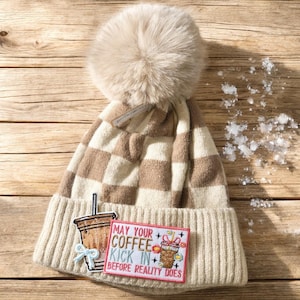 May include: A beige and brown checked beanie hat with a large, fluffy pom-pom. The hat has a ribbed cuff and a patch with a coffee cup graphic and the text "MAY YOUR COFFEE KICK IN BEFORE REALITY DOES."