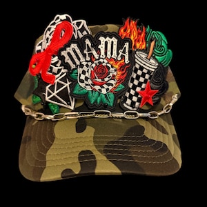 May include: A camouflage baseball cap featuring a silver chain across the brim and various embroidered patches including a red ribbon, a diamond, a rose with a checkered pattern, flames, a checkered cup, and the word MAMA in white lettering.