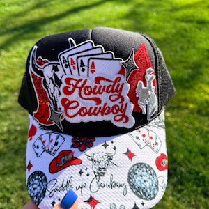 May include: Black and white trucker hat with a mesh back and a curved brim. The hat features a patch with the words "Howdy Cowboy" in red script, playing cards, a bull skull, and other western-themed graphics.