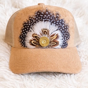 May include: Tan trucker hat with a mesh back and a suede front. The front of the hat is decorated with a cluster of feathers in white, brown, and black, and a gold-colored Winchester bullet casing.