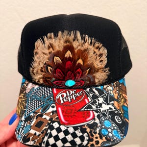 May include: Black trucker hat with a patterned brim featuring a Dr. Pepper can graphic. The hat has a large feather and turquoise stone embellishment. The brim has a mix of patterns including leopard print and checkered designs.