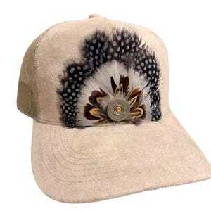 May include: Tan baseball cap with a mesh back and a suede-like front. The cap is decorated with a cluster of feathers in black, white, and brown, and a silver button. The hat is a stylish accessory.