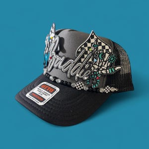 May include: A black trucker hat with a gray front panel and a mesh back. The hat features the word "Baddie" in silver script, embellished with decorative elements, including a checkered flag design and a skeleton hand.