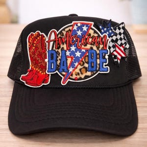 May include: Black trucker hat with a mesh back and a curved brim. The front features an embroidered patch with the words "American Babe" in red, white, and blue, along with a red cowboy boot, a lightning bolt, and stars.