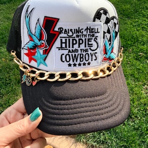 May include: Black and white trucker hat with a mesh back. The front features a patch with the text "Raising Hell with the Hippies and the Cowboys" and a lightning bolt. A gold chain with charms adorns the brim.