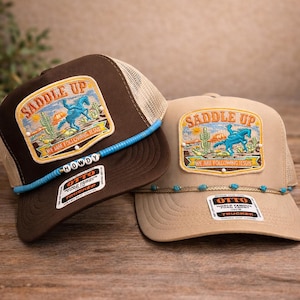 May include: Two trucker hats with a brown and tan color scheme. Each hat features a patch with a cowboy riding a horse, the words "SADDLE UP" and "WE ARE FOLLOWING JESUS". The brown hat has the word "HOWDY" on the brim.
