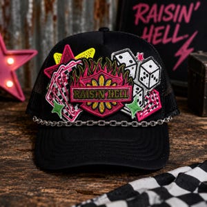 May include: A black trucker hat featuring a silver chain across the brim and embroidered patches including playing cards, dice, a yellow star, and a central shield emblem with flames and the text RAISIN' HELL in green and pink embroidery.