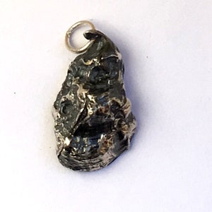 Könnte beinhalten: Ein silberner Anhänger in Form einer kleinen, detaillierten, strukturierten und abstrakten Figur. Der Anhänger hat eine Öse oben zum Anbringen an einer Halskette oder einem Armband.