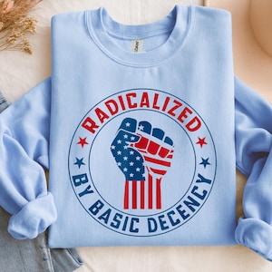 Op de afbeelding: Een lichtblauw sweatshirt met de tekst "RADICALIZED BY BASIC DECENCY" rond een vuistontwerp in rood, wit en blauw. Het ontwerp bevat de Amerikaanse vlag. Het sweatshirt is op een wit oppervlak gevouwen.