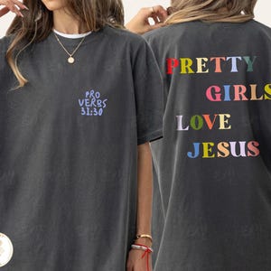 May include: A charcoal gray short-sleeve t-shirt featuring the text "PRO VERBS 31:30" in light blue on the front chest. The back displays the colorful text "PRETTY GIRLS LOVE JESUS" in red, pink, yellow, light blue, and lavender lettering.