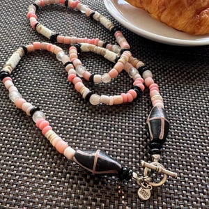 May include: A beaded necklace featuring pink, white, and black beads. The necklace has two large black and pink decorative beads and a silver toggle clasp. A croissant rests on a white plate in the background.