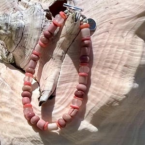 May include: A pink beaded bracelet with a silver clasp. The bracelet features a variety of pink beads, including round and cylindrical shapes, and is displayed on a light-colored surface.