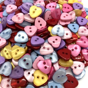 May include: A collection of heart-shaped buttons in a variety of colours, such as red, pink, yellow, purple, and blue. Each button has two holes for sewing. The buttons are made of a glossy material.