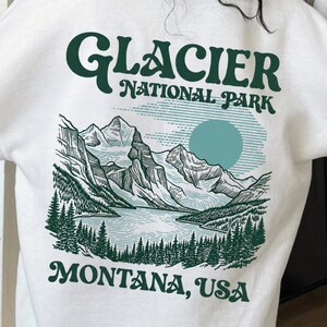 May include: A white long-sleeve sweatshirt featuring a dark green graphic on the back. The design displays a mountain landscape, a lake, and pine trees with the text GLACIER NATIONAL PARK and MONTANA, USA in a dark green serif font.