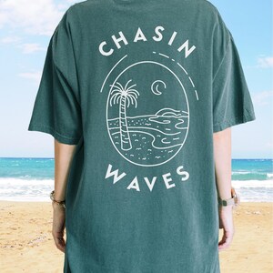 以下が含まれることがあります： 背面に白いグラフィックが施されたダークグリーンの半袖Tシャツ。デザインには「CHASIN WAVES」という文字が含まれており、ヤシの木、三日月、砂浜の海岸線に沿った海の波が描かれた楕円形のフレームを囲んでいます。