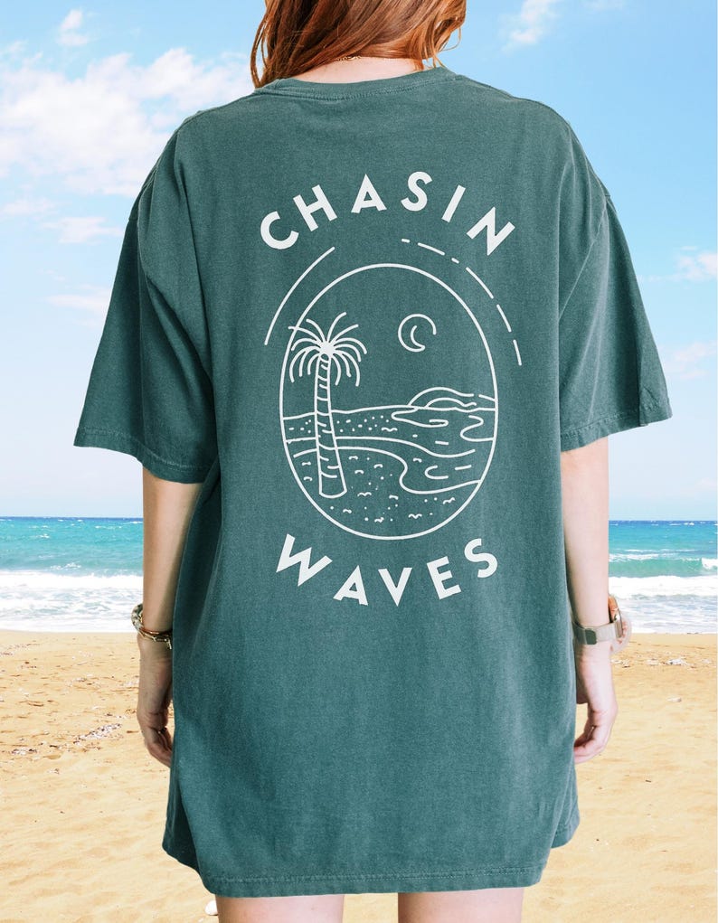 以下が含まれることがあります： 背面に白いグラフィックが施されたダークグリーンの半袖Tシャツ。デザインには「CHASIN WAVES」という文字が含まれており、ヤシの木、三日月、砂浜の海岸線に沿った海の波が描かれた楕円形のフレームを囲んでいます。