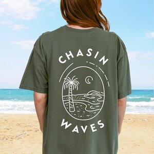 以下が含まれることがあります： 背面に白いグラフィックがプリントされたオリーブグリーンの半袖Tシャツ。デザインは円形の輪郭の中にヤシの木、三日月、海の波が描かれており、その周囲に「CHASIN WAVES」というテキストがアーチ状に配置されています。