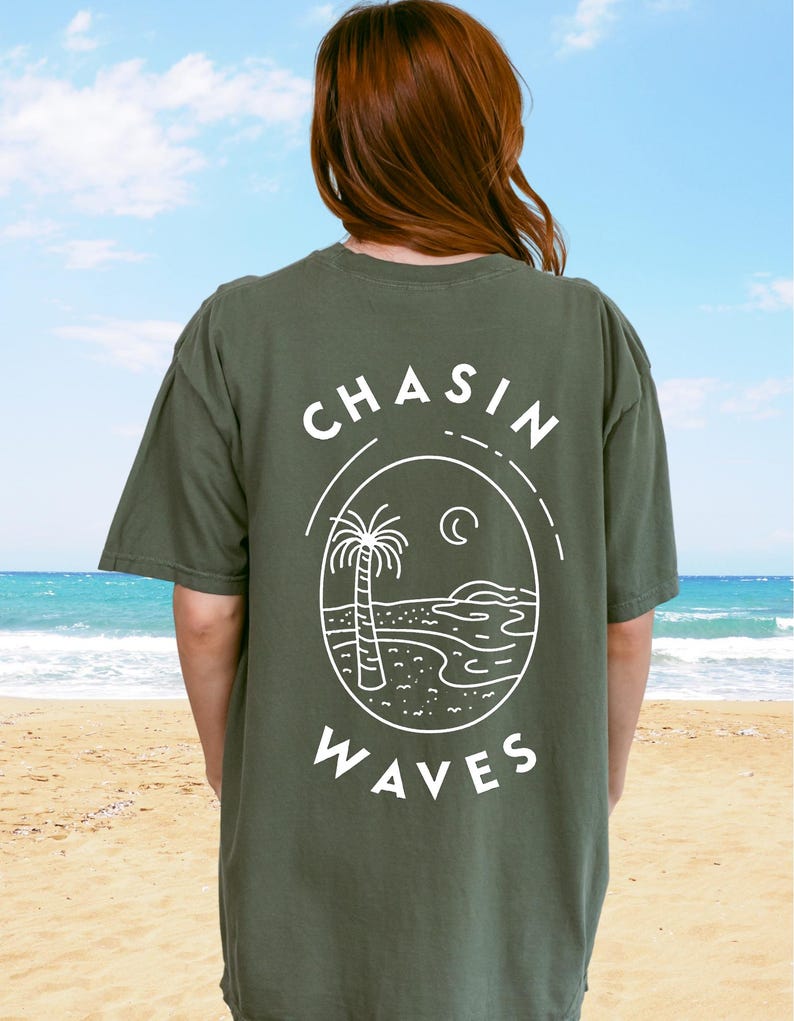 以下が含まれることがあります： 背面に白いグラフィックがプリントされたオリーブグリーンの半袖Tシャツ。デザインは円形の輪郭の中にヤシの木、三日月、海の波が描かれており、その周囲に「CHASIN WAVES」というテキストがアーチ状に配置されています。