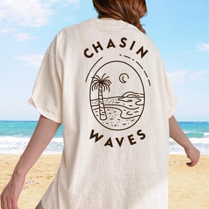 以下が含まれることがあります： オフホワイトの半袖Tシャツで、背面にダークブラウンの線画グラフィックが施されています。デザインは、ヤシの木、海の波、三日月が描かれた楕円形のフレームで構成されており、周囲に「CHASIN WAVES」というテキストがプリントされています。