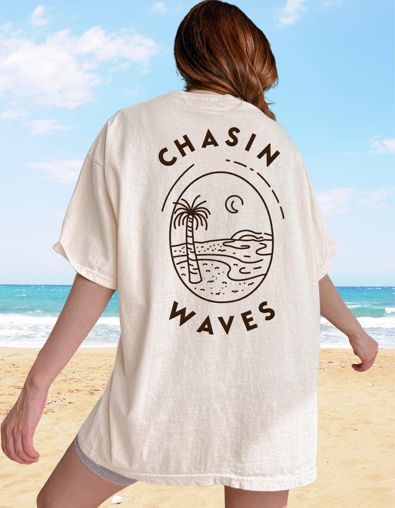 以下が含まれることがあります： オフホワイトの半袖Tシャツで、背面にダークブラウンの線画グラフィックが施されています。デザインは、ヤシの木、海の波、三日月が描かれた楕円形のフレームで構成されており、周囲に「CHASIN WAVES」というテキストがプリントされています。