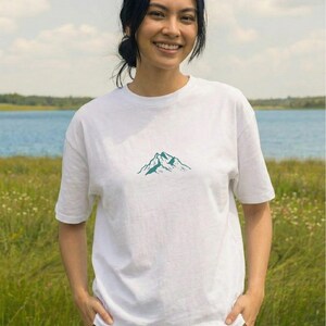 May include: White t-shirt with a teal mountain graphic. The shirt is made of a soft material and has short sleeves. The person is smiling and standing in a field with a lake in the background.