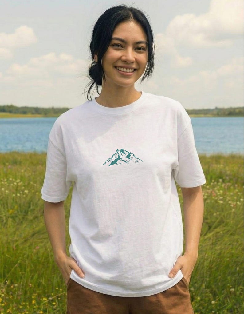 May include: White t-shirt with a teal mountain graphic. The shirt is made of a soft material and has short sleeves. The person is smiling and standing in a field with a lake in the background.