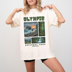 May include: Cream-colored t-shirt featuring the text "OLYMPIC" in green block letters above four nature scene illustrations. The illustrations depict a coastal landscape, a forest, and wildlife. The words "NATIONAL PARK" and "WASHINGTON" are also printed on the shirt.