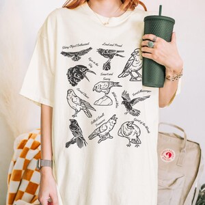 May include: Cream-colored t-shirt featuring black illustrations of various birds with phrases like "Shiny Object Enthusiast" and "Collect Small Treasures." A dark green tumbler is held in the hand.