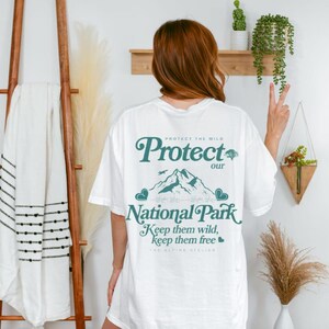 May include: White t-shirt with a teal graphic that reads "Protect our National Park." The design includes mountains, trees, and the text "Keep them wild, keep them free." The shirt is worn by a person.
