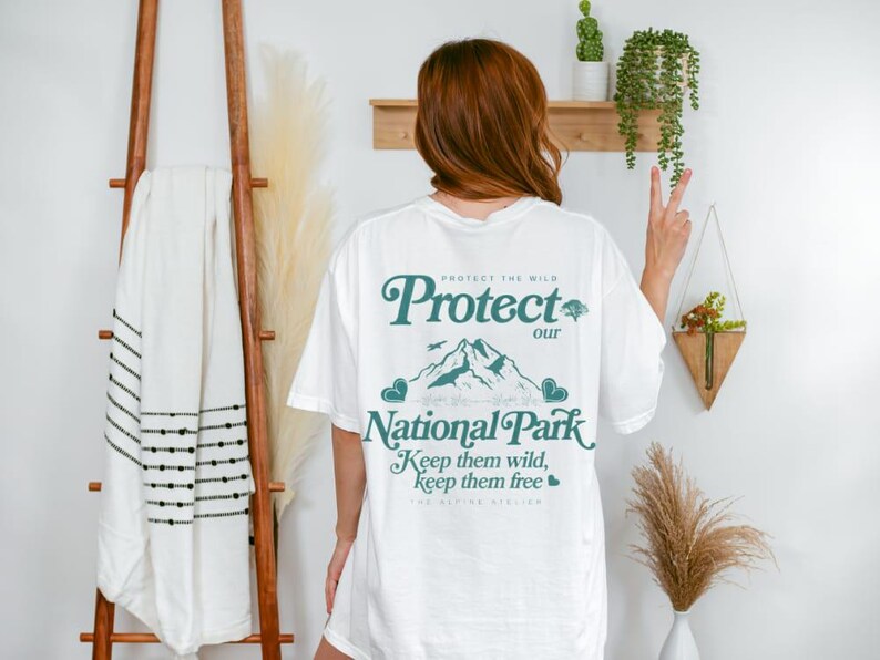 May include: White t-shirt with a teal graphic that reads "Protect our National Park." The design includes mountains, trees, and the text "Keep them wild, keep them free." The shirt is worn by a person.
