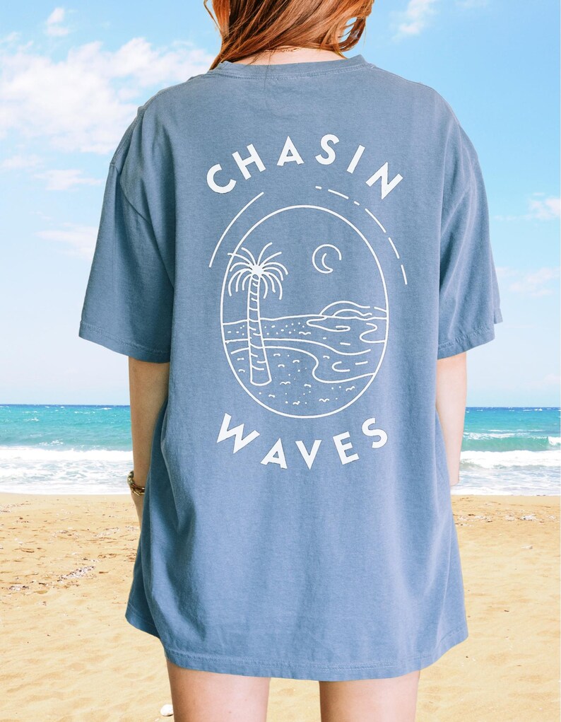 以下が含まれることがあります： 背面に白いグラフィックがプリントされた、ダスティブルーの半袖Tシャツ。楕円形のフレームの中にヤシの木、海の波、三日月が描かれており、その上下に「CHASIN WAVES」というテキストがアーチ状に配置されています。