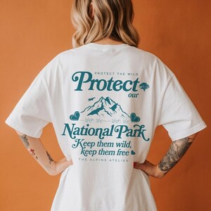 May include: White t-shirt with a teal graphic that reads "Protect our National Park." The design includes mountain illustrations, birds, and the text "Keep them wild, keep them free." The Alpine Atelier is also printed on the shirt.