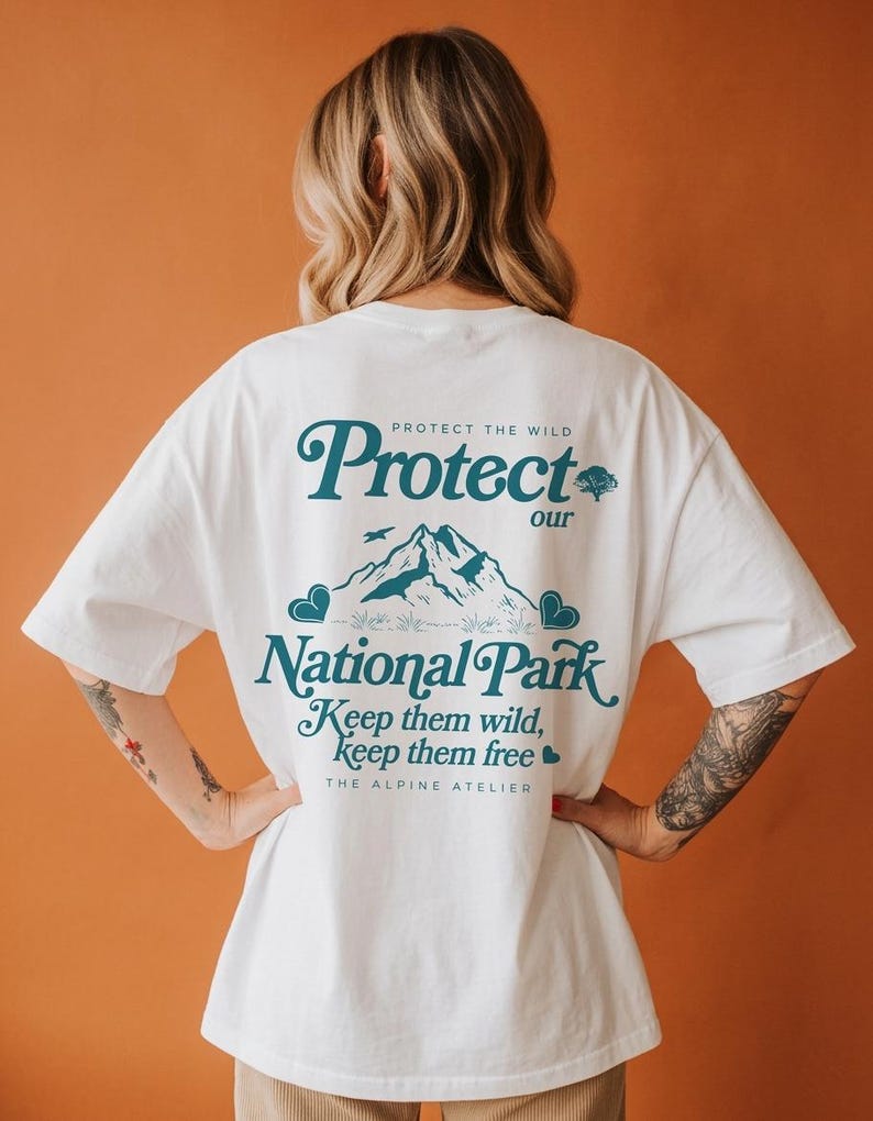 May include: White t-shirt with a teal graphic that reads "Protect our National Park." The design includes mountain illustrations, birds, and the text "Keep them wild, keep them free." The Alpine Atelier is also printed on the shirt.