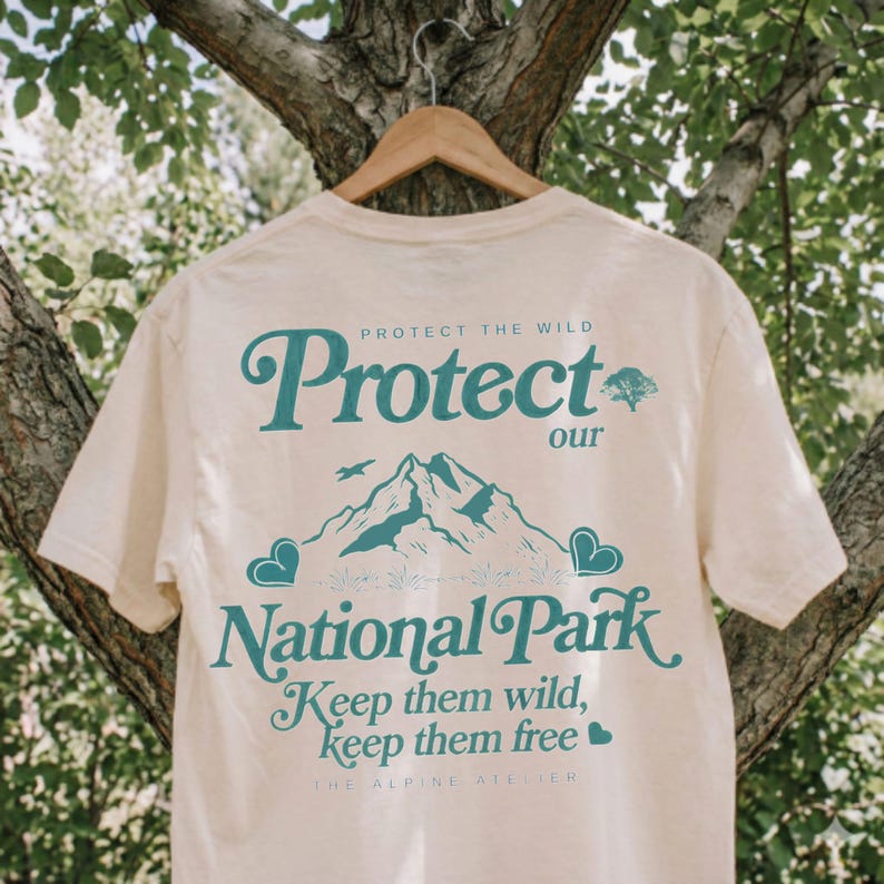 May include: Cream-colored t-shirt with teal text and graphics. The shirt features the words "Protect our National Park" and "Keep them wild, keep them free." A mountain range and heart-shaped graphics are also included.