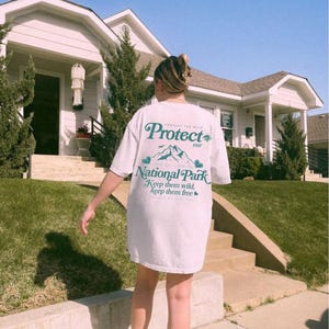 May include: White t-shirt with the text "PROTECT our NATIONAL PARK Keep them wild, Keep them free" in teal. The shirt features a mountain graphic. The person is standing on a sidewalk in front of a house.