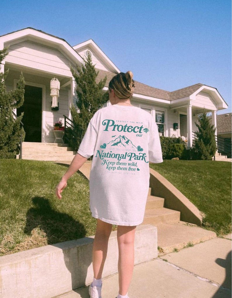 May include: White t-shirt with the text "PROTECT our NATIONAL PARK Keep them wild, Keep them free" in teal. The shirt features a mountain graphic. The person is standing on a sidewalk in front of a house.