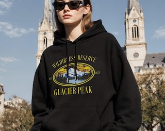 May include: Black hoodie with a front pocket and drawstring hood. The hoodie features a yellow and white graphic with the text "Wilderness Reserve Glacier Peak" and the year "1885". The person is wearing black sunglasses.
