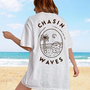 以下が含まれることがあります： 背面にダークブラウンのグラフィックがプリントされた白い半袖Tシャツ。デザインは、ヤシの木、海の波、三日月が描かれたビーチシーンの円形線画で、その周囲に太字でCHASIN WAVESというテキストが配置されています。